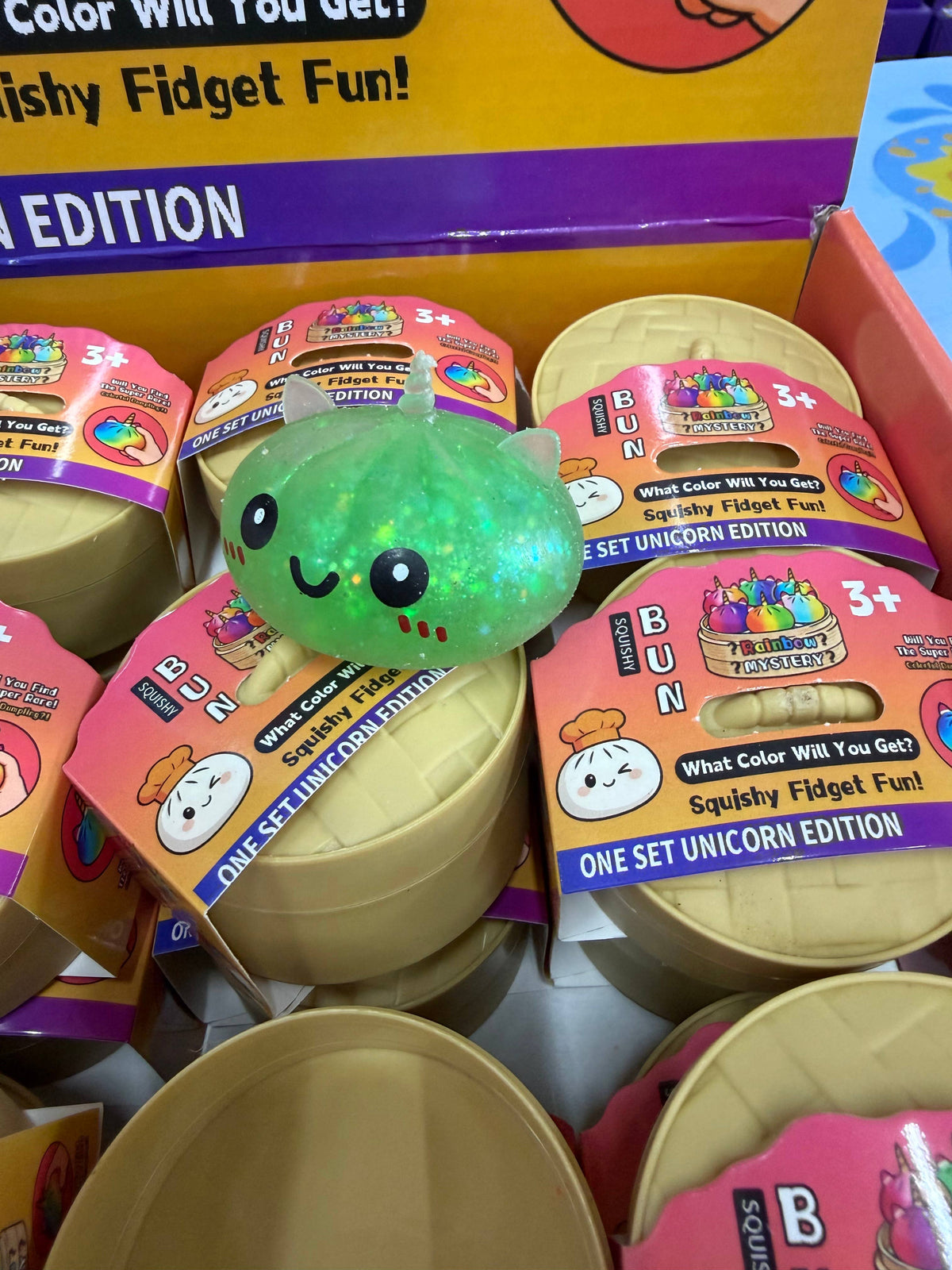 A green, smiling MAKLY CO Squishy Dumpling—Small Glitter Unicorn Metallic Colors Slow (Single)—sits atop sealed round containers labeled Squishy Fidget Fun! One Set Unicorn Edition in a store display for children ages 3 and up, offering a delightful stress relief toy experience.