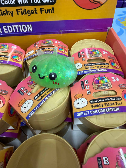 A green, smiling MAKLY CO Squishy Dumpling—Small Glitter Unicorn Metallic Colors Slow (Single)—sits atop sealed round containers labeled Squishy Fidget Fun! One Set Unicorn Edition in a store display for children ages 3 and up, offering a delightful stress relief toy experience.