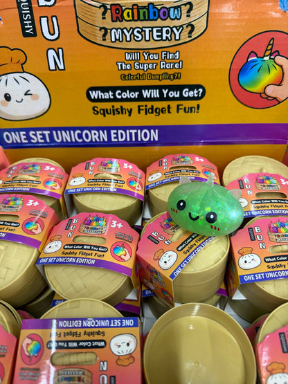 A display of MAKLY CO Squishy Dumpling Small Glitter Unicorn Metallic Colors Slow fidget toys features eye-catching gold capsule packaging and a green, dumpling-shaped squishy with a smiley face on top. Vibrant signage highlights the unicorn edition from this stress relief toy collection.