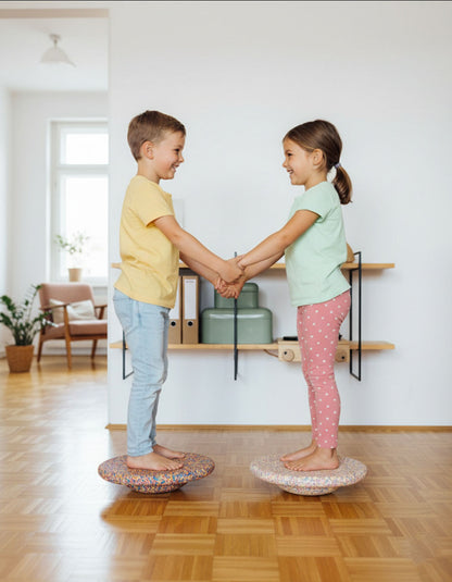 Two children stand facing each other on Okie Soft Play Store’s Rainbow Balance Board for Kids, holding hands and smiling as they play in a bright living room with wooden floors and minimal decor, enjoying this wobble board designed for active play.