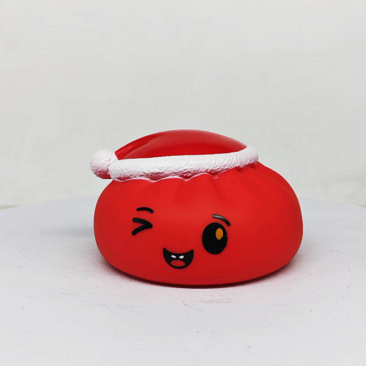 The Christmas Bun Squeaky Toy by Okie Soft Play is a red, round toy featuring a winking cartoon face and a Santa hat, designed to resemble a playful holiday bun. It is shown against a plain white background.